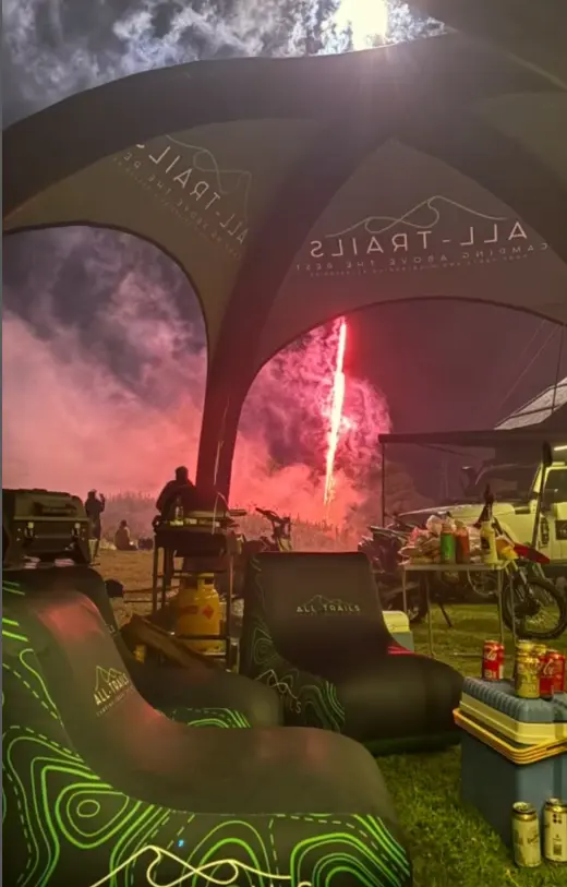 A cozy, evening atmosphere under a branded "ALL TRAILS" inflatable tent, with people relaxing, drinks on a table, and a bright red flare or firework display illuminating the sky in the background.